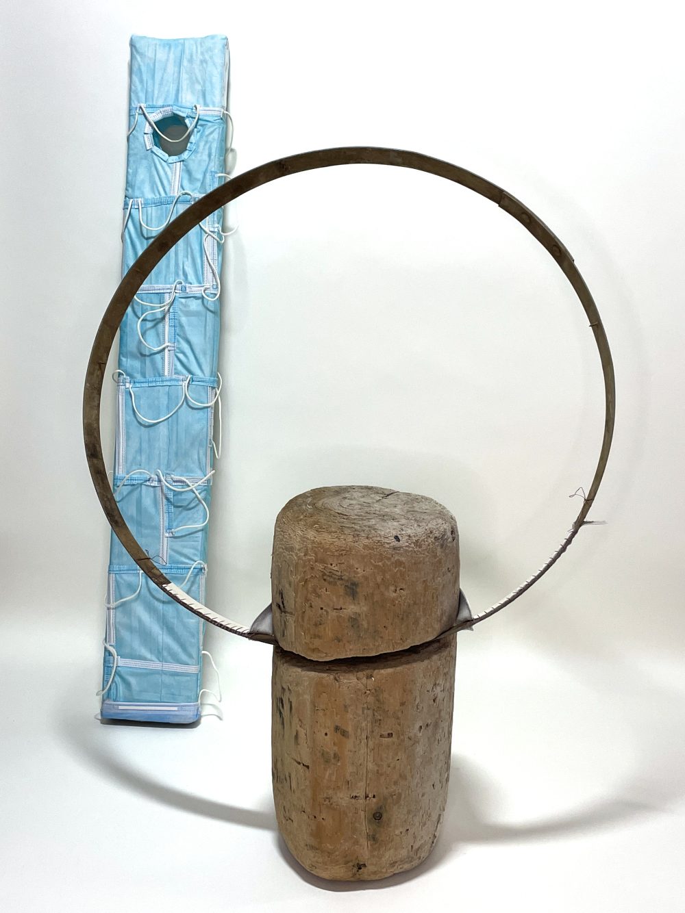 Two sculptures; one of a standing piece of wood with a metal ring and the other behind it, a tall structure covered in blue masks.