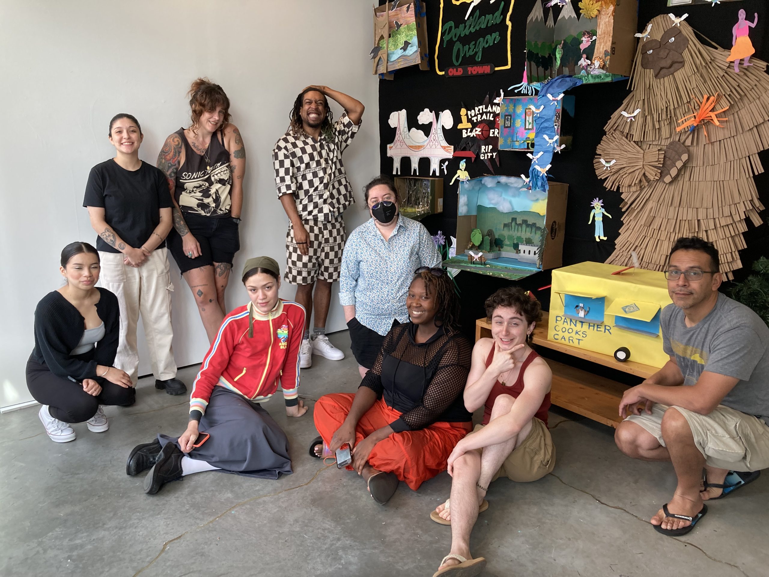 Nine students pose in front of their collaborative work with paper materials.