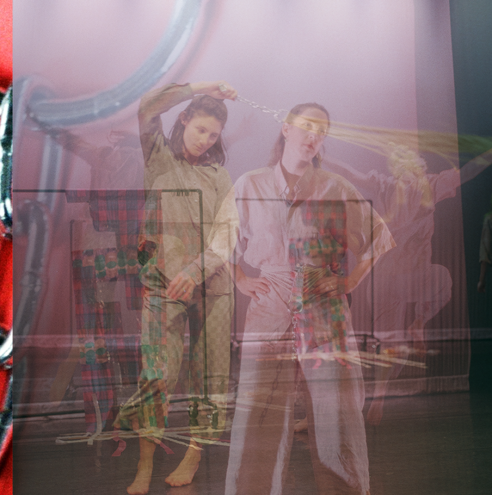 A layered image of Hannah and Emily. Emily is swinging a rope attached to a chain, taken from a hammock. Hannah is wearing the same object around their waist. The two are layered on one another, and ghostly images of them jumping onstage are in the background. Behind it all, and peeking through the left side, is the hammock chain on bright pink fabric. This image is extremely collaged.