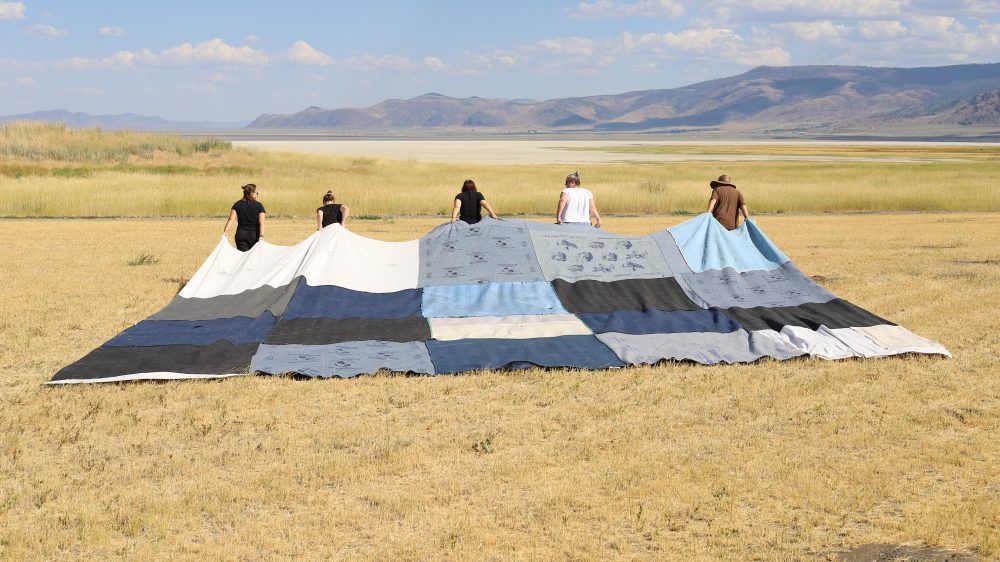 (promotional image description) image of five people carrying a large quilt in a desert near the mountains