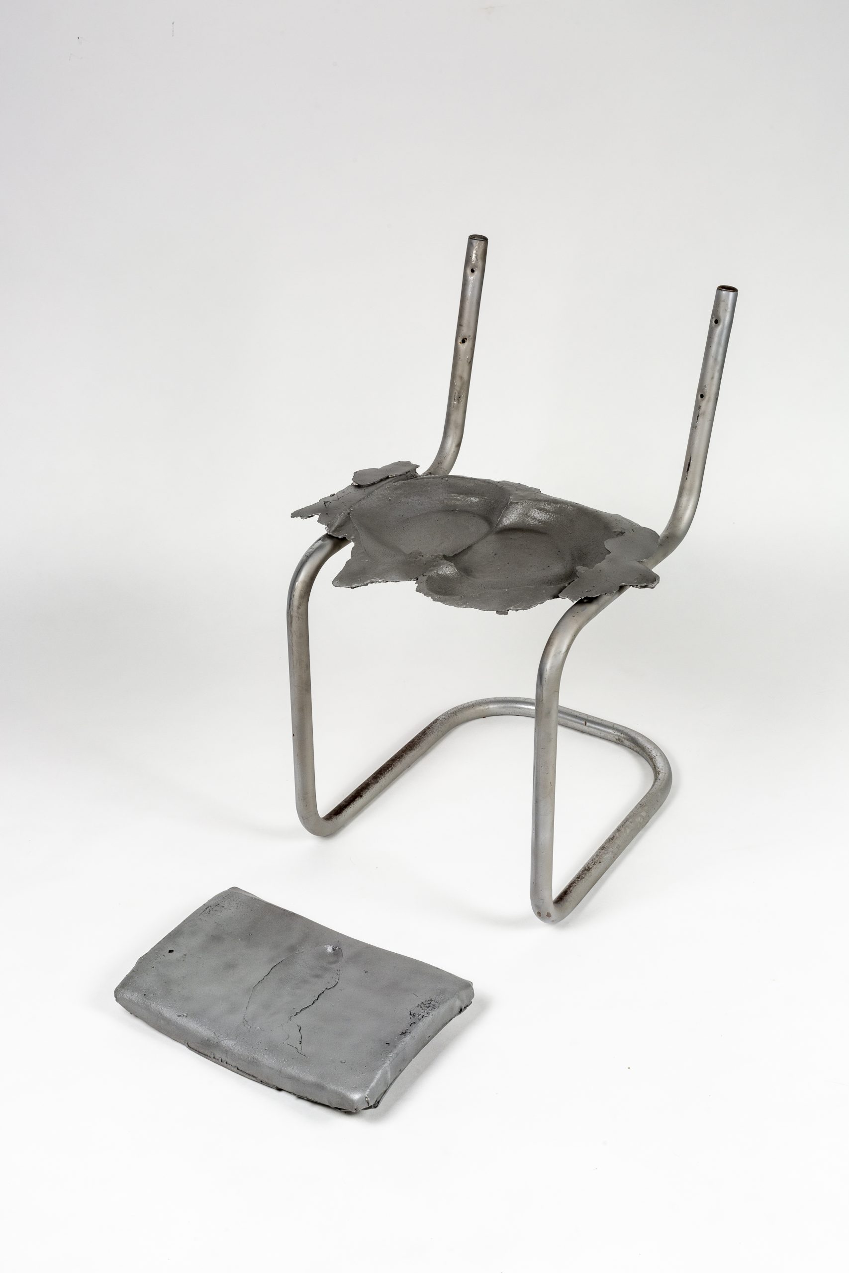 Tubular steel chair frame with an impression in the seat and a clay piece with an impression on the ground in front of it.