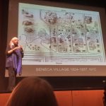 Scholar speaking in front of an audience, with a drawing of the Seneca Village projected on the screen in the background.