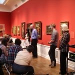 Students standing in front of paintings with an audience sitting on chairs in front of them.
