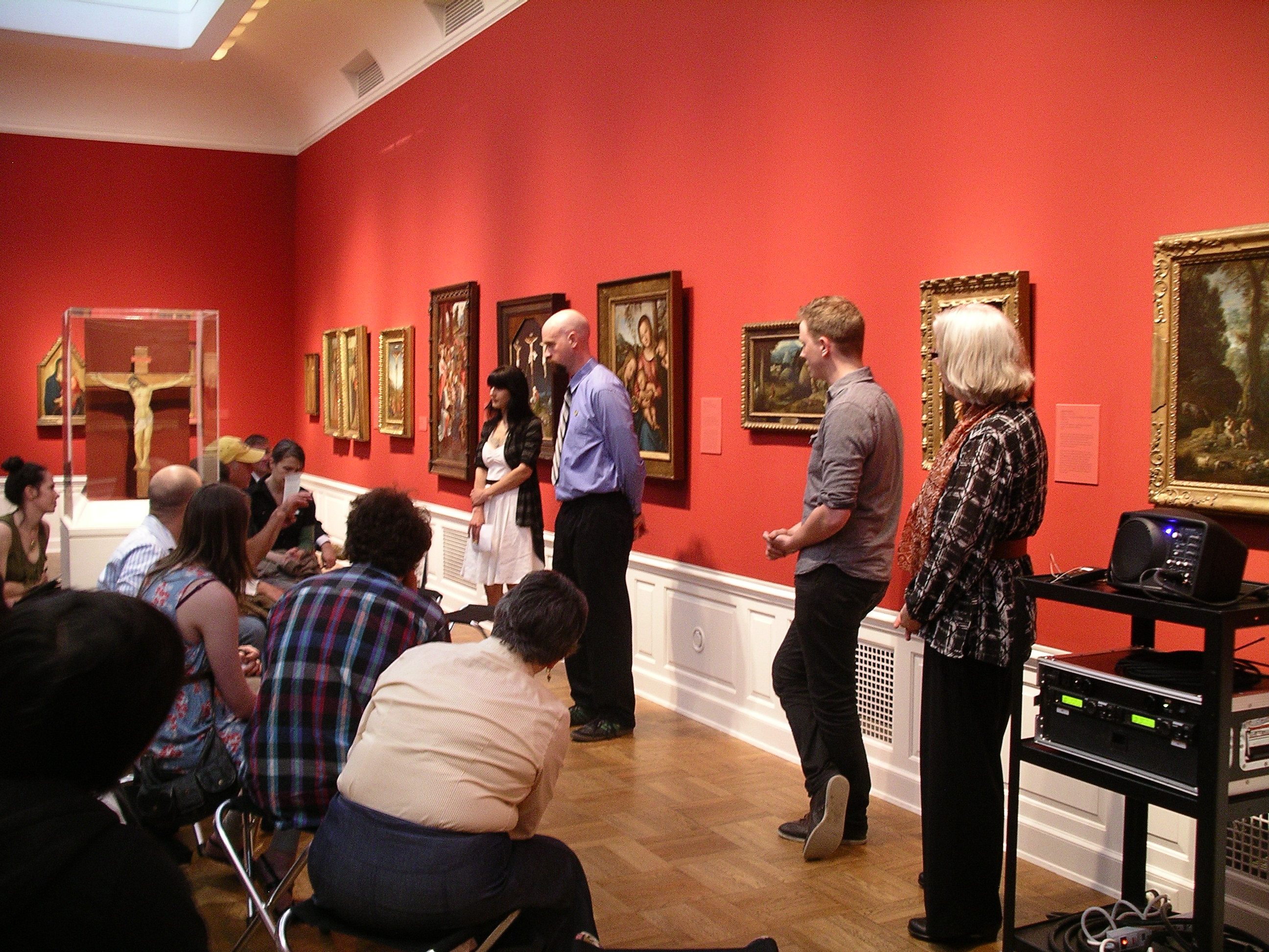 Students standing in front of paintings with an audience sitting on chairs in front of them.