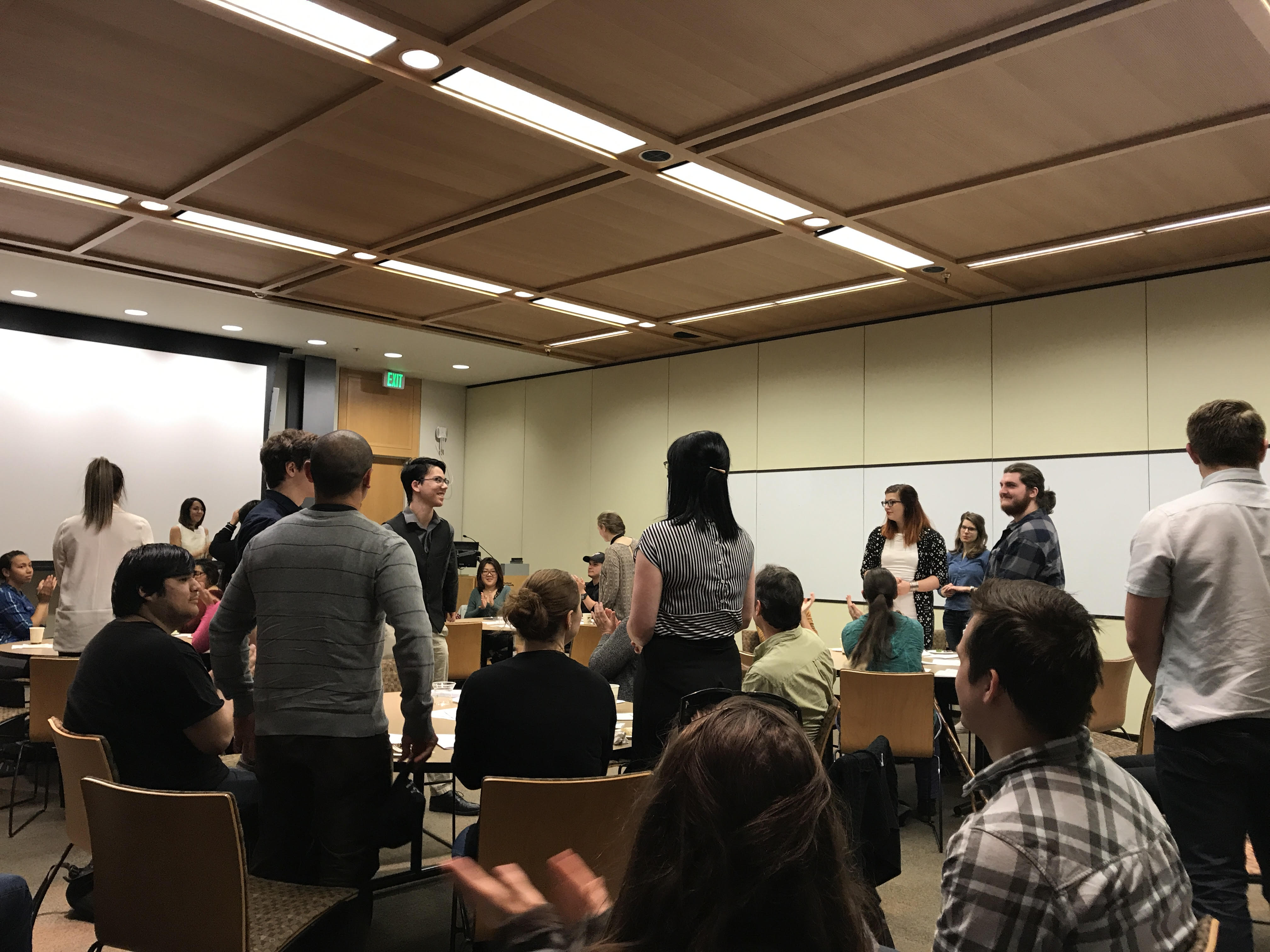 Students standing around tables and others clapping for them.