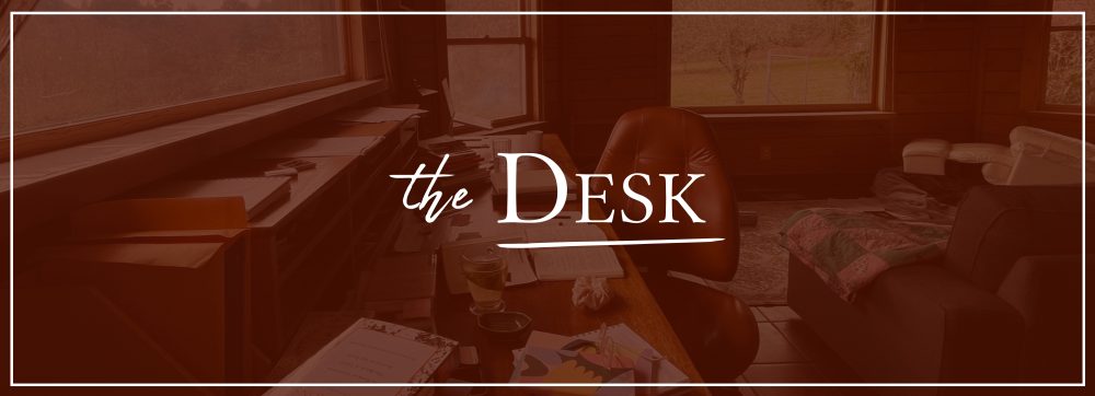 A red photo of a desk inside the Writers House overlaid with white text that says "The Desk"