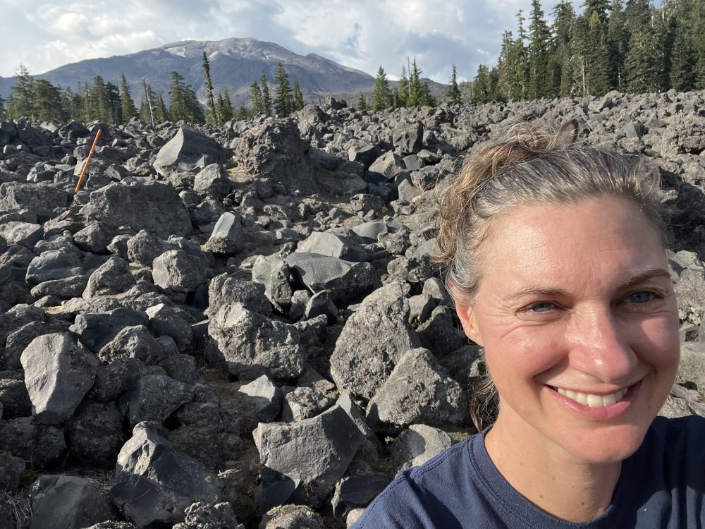 White woman with brown hair and blue eyes smiling in front of grey rocks, green pine trees, and a mountain.