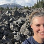 White woman with brown hair and blue eyes smiling in front of grey rocks, green pine trees, and a mountain.