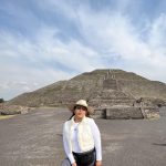 Mexican woman with brown hair, a white hat, white shirt and vest, and grey pants standing in front of a Mayan pyramid.