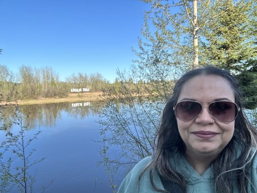 Woman with brown skin and brown hair, wearing sunglasses, standing in front of water and trees. 