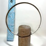 Two sculptures; one of a standing piece of wood with a metal ring and the other behind it, a tall structure covered in blue masks.