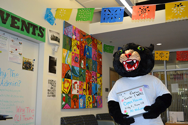 Poppy the Panther standing in front of a mural holding a sign that reads wrc and mc open house