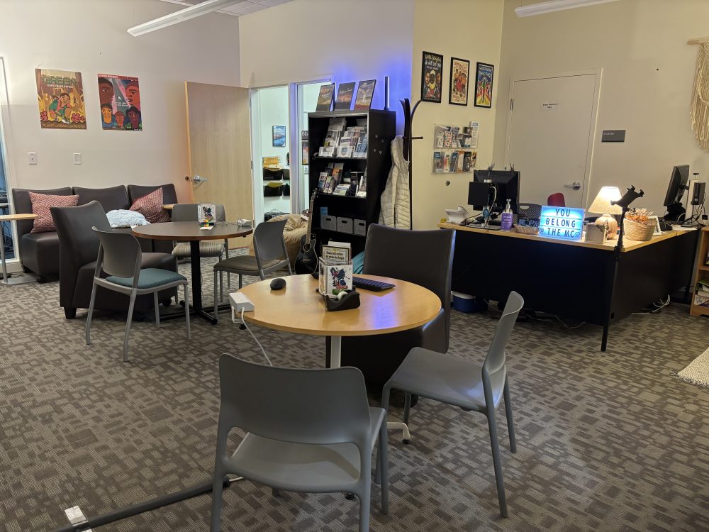 A picture of inside the Southeast Multicultural Center. There are two small round tables with chairs placed around them, a small brown couch with pillows, a bookshelf with flyers and snacks, and a front desk with a sign that reads "You belong at the MC"