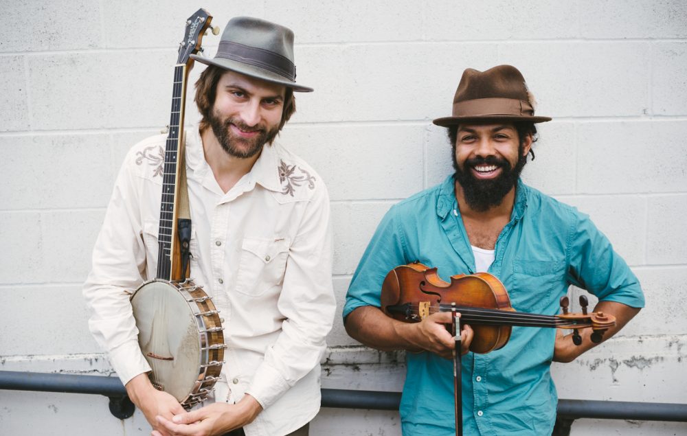 two men standing against wall holding banjo and fiddle