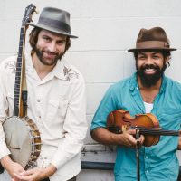 two men standing against wall holding banjo and fiddle
