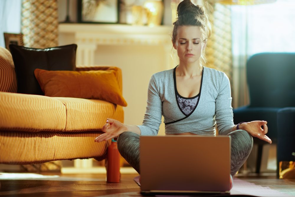 relaxed healthy sports woman in fitness clothes at modern home meditating using online streaming yoga site in laptop.