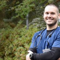 Nursing student standing outside with their arms folded and wearing scrubs, smiling into the camera