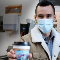 Hector Mejía Zamora in a coffee shop wearing a mask and holding a cup of coffee up to the camera