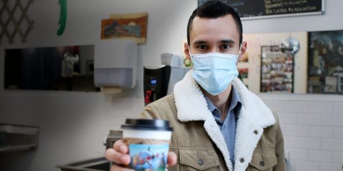 Hector Mejía Zamora in a coffee shop wearing a mask and holding a cup of coffee up to the camera