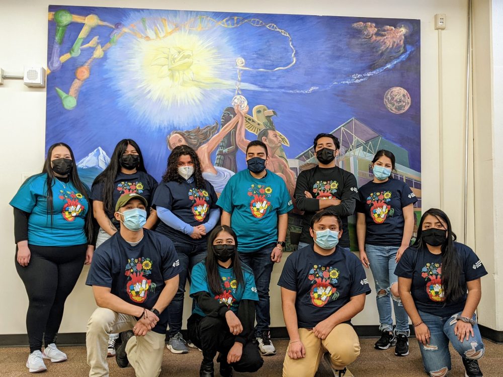 group of students wearing protective face masks pose in front of a colorful painting