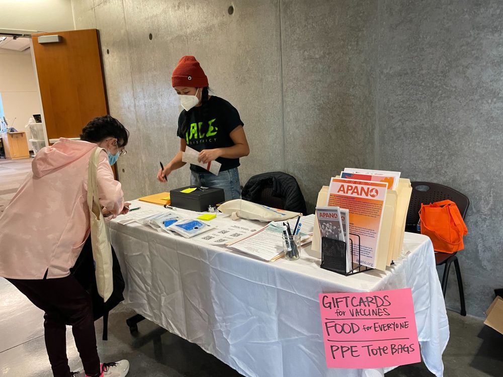 Table with resources and pamphlets. Person standing behind the table assisting another person looking at the information.