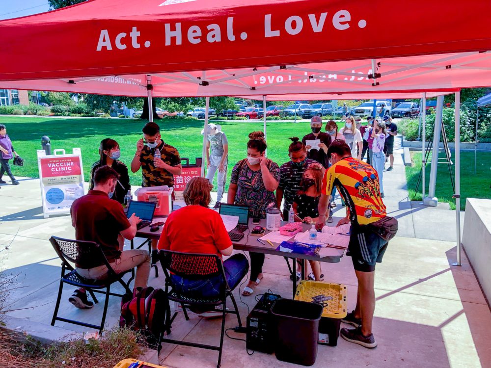people standing in line and registering for their vaccine; tent with the words Act.Heal.Love