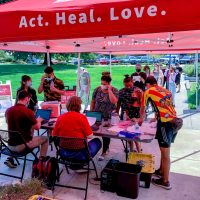 people standing in line and registering for their vaccine; tent with the words Act.Heal.Love