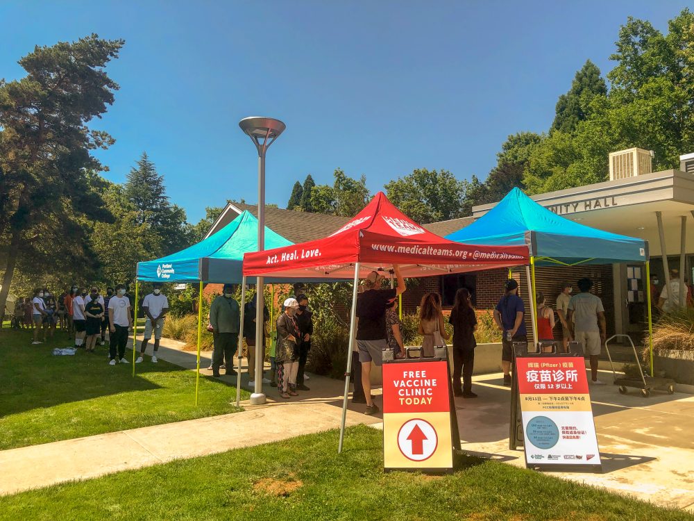 people standing in line outdoors on a sunny day waiting to get their vaccine, sandwhich boards with information in English and Chinese