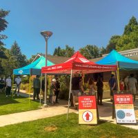 people standing in line outdoors on a sunny day waiting to get their vaccine, sandwhich boards with information in English and Chinese