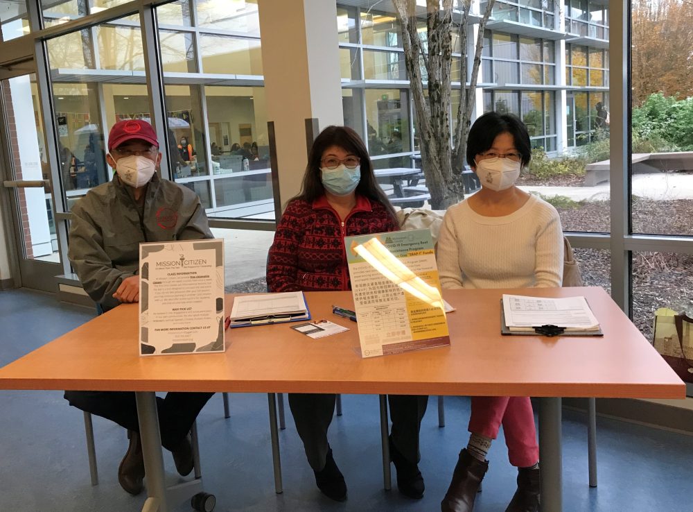 3 people sitting at a table wearing masks with informational materials