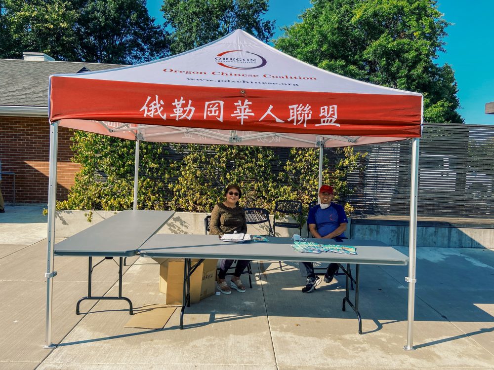 Two people sitting under a tent with the Oregon Chinese Coalition logo and other words in Chinese