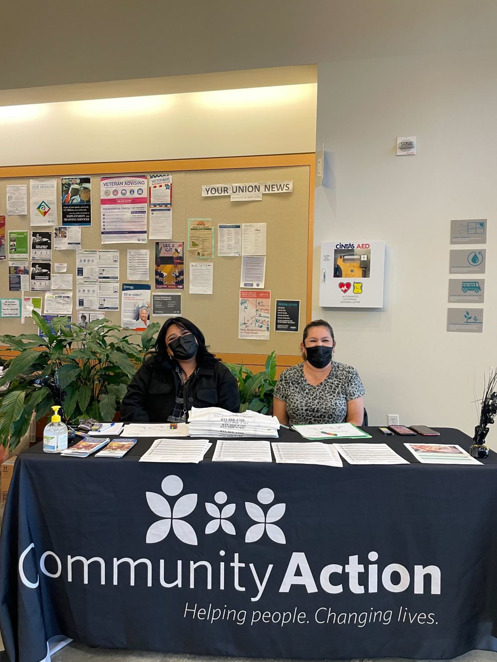 Table with two people sitting behind it providing flyers and information. The tablecloth reads Community Action - Helping people. Changing Lives.