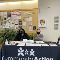 Table with two people sitting behind it providing flyers and information. The tablecloth reads Community Action - Helping people. Changing Lives.