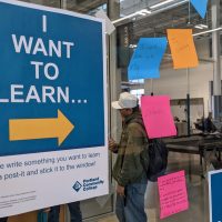 Sign reading "I Want to Learn" with arrow pointing to glass wall and brainstorm post-it notes