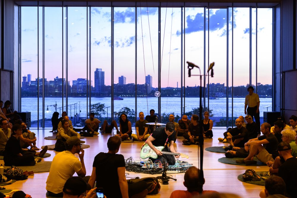 City in background of crowd gathered around sound musician at Whitney Museum.