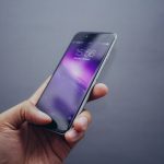 A medium tone hand holds a smartphone with their thumb on the home button