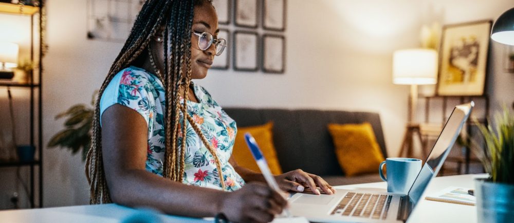 A woman with long braids and glasses sits at a desk working on a laptop. She is holding a pen and writing in a notebook, with a coffee mug beside her. The room is softly lit with a lamp and decorated with yellow pillows and framed art in the background.
