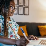 A woman with long braids and glasses sits at a desk working on a laptop. She is holding a pen and writing in a notebook, with a coffee mug beside her. The room is softly lit with a lamp and decorated with yellow pillows and framed art in the background.