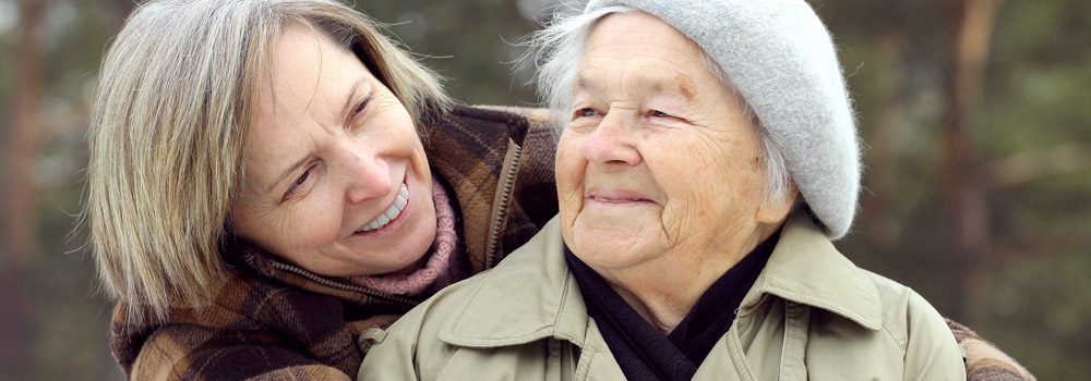 Aging adult mother looks up as she is hugged by her adult daughter.