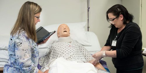  An instructor coaches a female student as she practices entry level CNA skills on healthcare mannequin in bed