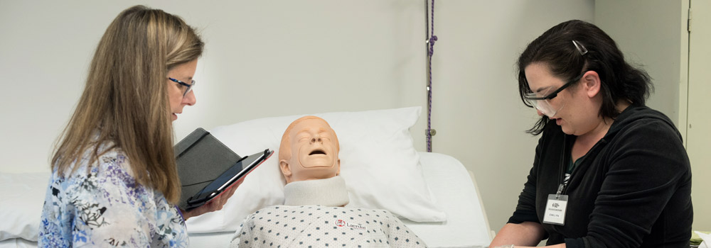 An instructor coaches a female student as she practices entry level CNA skills on healthcare mannequin in bed