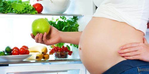 Image of pregnant woman in front of a refrigerator holding an apple.