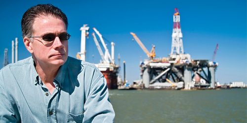 Man sitting in front of ocean with industrial rig in background
