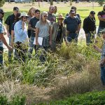 A group of adults stands outdoors in a landscaped area, listening to an instructor - David Sandrock, who is pointing out plants and grasses during a hands-on landscaping or horticulture training session in a park-like setting.