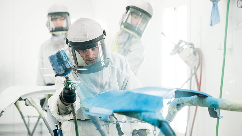 Students wearing protective equpiment spray painting a car door blue in a white room