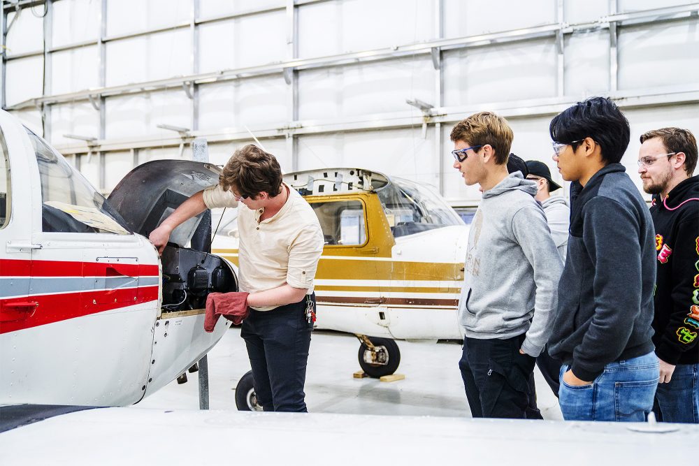 Students watch an instructor demonstrate working on an airplane engine in the hangar
