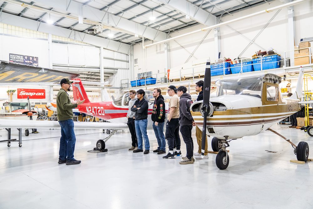 Students listening to an instructor among the airplanes in the aviation hangar