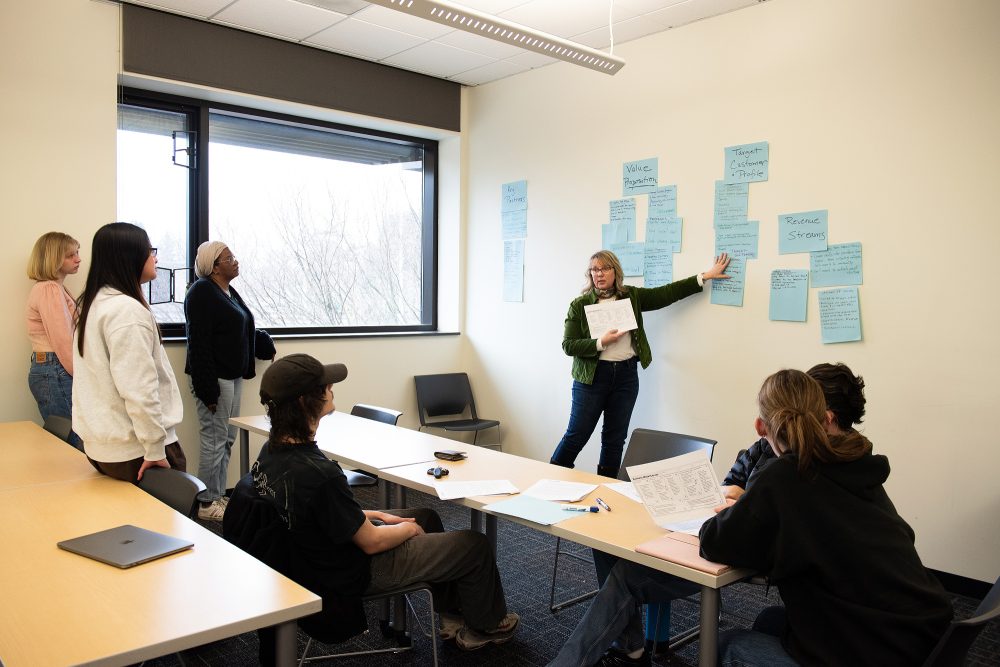 Instructor pointing at a wall of sticky notes with words like "value proposition" and "target customer"
