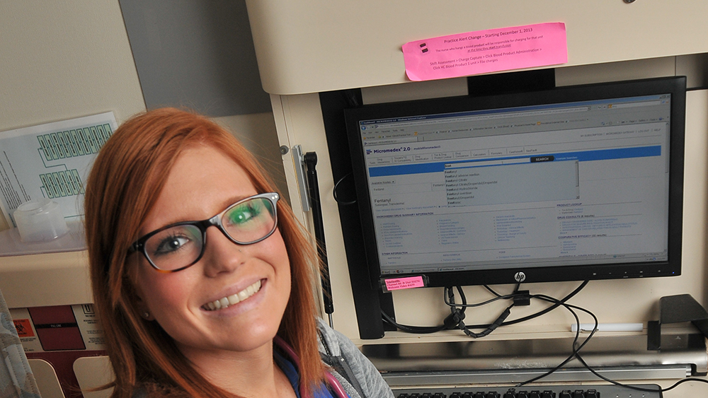 Student in medical attire standing in front of a computer in a medical office