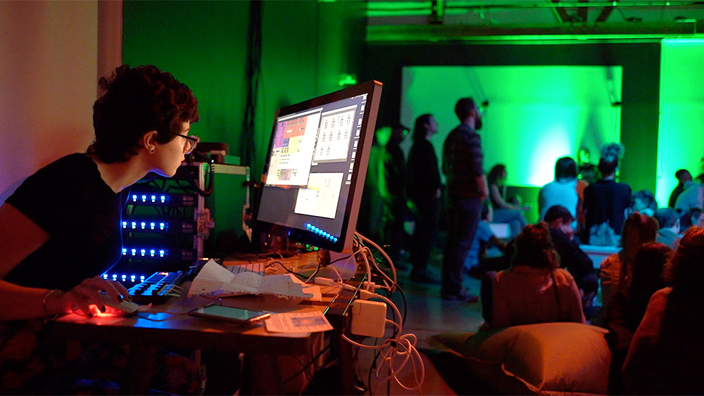 Student working on a computer with audio equipment while other students stand by a greenscreen in the background